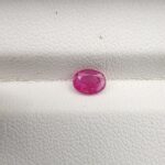 A natural 0.50-carat pinkish-red ruby (Yaqoot) from Mozambique, displaying a faceted oval cut with a distinct semi-transparent crystal structure and natural internal inclusions, resting on a textured white surface.