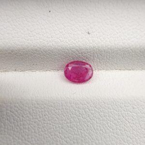 A natural 0.50-carat pinkish-red ruby (Yaqoot) from Mozambique, displaying a faceted oval cut with a distinct semi-transparent crystal structure and natural internal inclusions, resting on a textured white surface.