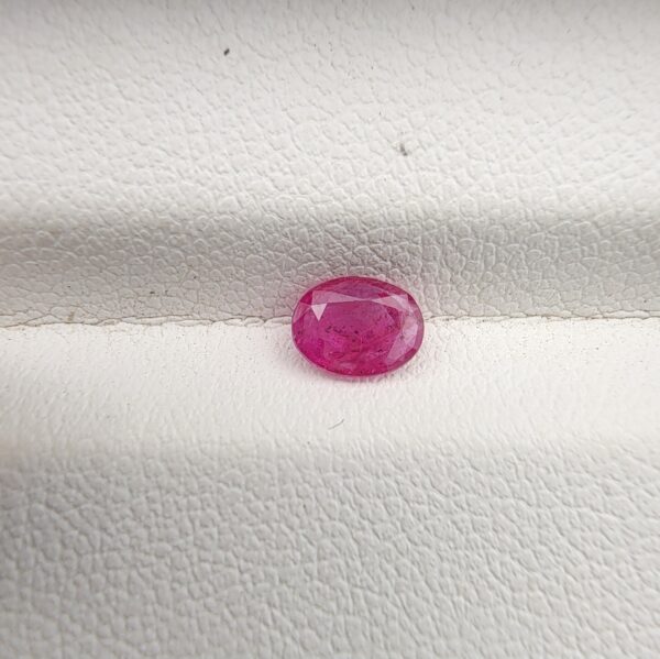 A natural 0.50-carat pinkish-red ruby (Yaqoot) from Mozambique, displaying a faceted oval cut with a distinct semi-transparent crystal structure and natural internal inclusions, resting on a textured white surface.