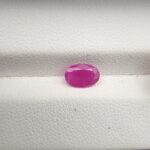 A natural 1.03-carat pinkish-red ruby (Yaqoot) from Mozambique, displaying a faceted oval cut with a distinct translucent crystal structure and natural internal inclusions, resting on a textured white surface.