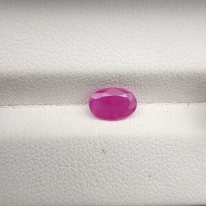 A natural 1.03-carat pinkish-red ruby (Yaqoot) from Mozambique, displaying a faceted oval cut with a distinct translucent crystal structure and natural internal inclusions, resting on a textured white surface.