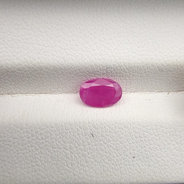 A natural 1.03-carat pinkish-red ruby (Yaqoot) from Mozambique, displaying a faceted oval cut with a distinct translucent crystal structure and natural internal inclusions, resting on a textured white surface.
