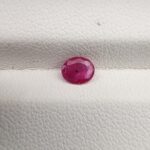 A natural 0.48-carat pinkish-red ruby (Yaqoot) from Mozambique, displaying a faceted oval cut with a distinct semi-transparent crystal structure and minor natural pinpoint inclusions, resting on a textured white surface.