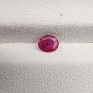A natural 0.48-carat pinkish-red ruby (Yaqoot) from Mozambique, displaying a faceted oval cut with a distinct semi-transparent crystal structure and minor natural pinpoint inclusions, resting on a textured white surface.