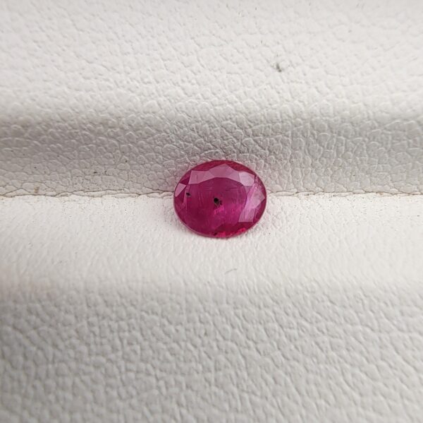 A natural 0.48-carat pinkish-red ruby (Yaqoot) from Mozambique, displaying a faceted oval cut with a distinct semi-transparent crystal structure and minor natural pinpoint inclusions, resting on a textured white surface.