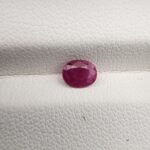 A natural 0.61-carat red ruby (Yaqoot) from Mozambique, displaying a faceted oval cut with a distinct translucent crystal structure and natural internal inclusions, resting on a textured white surface.