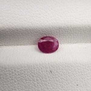 A natural 0.61-carat red ruby (Yaqoot) from Mozambique, displaying a faceted oval cut with a distinct translucent crystal structure and natural internal inclusions, resting on a textured white surface.