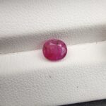 A natural 1.25-carat pinkish-red ruby (Yaqoot) from Mozambique, displaying a faceted oval cut with a distinct semi-transparent crystal structure and noticeable natural internal inclusion markers, resting on a textured white surface.