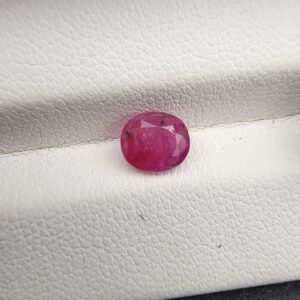 A natural 1.25-carat pinkish-red ruby (Yaqoot) from Mozambique, displaying a faceted oval cut with a distinct semi-transparent crystal structure and noticeable natural internal inclusion markers, resting on a textured white surface.