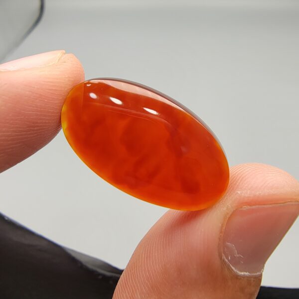 A premium 18.91-carat natural Yemeni Aqeeq agate gemstone with a polished oval cabochon cut, displaying a rich translucent orange-red color profile held securely in professional jeweler's tweezers.