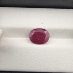 A premium 3.24-carat natural, unheated Mozambican ruby showing a faceted oval shape with a deep purplish-red translucent color profile, resting inside a textured white leather jewelry box liner.