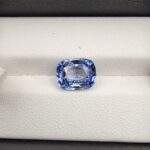​A premium 4.96-carat natural Ceylon blue sapphire gemstone displaying an elegant cushion cut with excellent symmetry and a deep royal blue color profile, resting inside a textured white leather jewelry box liner.
