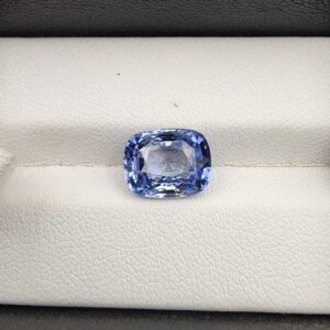 ​A premium 4.96-carat natural Ceylon blue sapphire gemstone displaying an elegant cushion cut with excellent symmetry and a deep royal blue color profile, resting inside a textured white leather jewelry box liner.