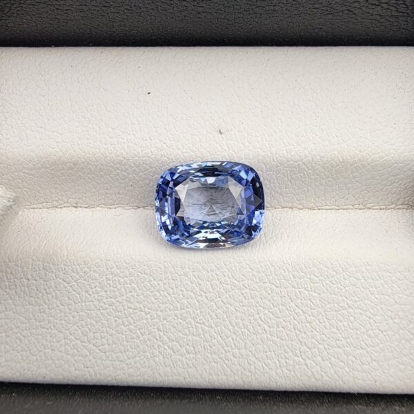 ​A premium 4.96-carat natural Ceylon blue sapphire gemstone displaying an elegant cushion cut with excellent symmetry and a deep royal blue color profile, resting inside a textured white leather jewelry box liner.
