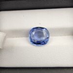 A premium 5.15-carat natural Ceylon blue sapphire gemstone displaying an elegant cushion cut with excellent symmetry and a deep royal blue color profile, resting inside a textured white leather jewelry box liner.