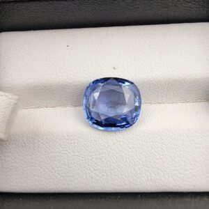 A premium 5.15-carat natural Ceylon blue sapphire gemstone displaying an elegant cushion cut with excellent symmetry and a deep royal blue color profile, resting inside a textured white leather jewelry box liner.