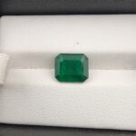 ​A premium 2.98-carat natural Zambian emerald gemstone displaying an elegant octagonal step cut with sharp parallel facet lines and a rich green transparency, resting inside a textured white leather jewelry box liner.