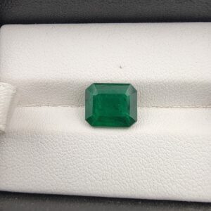 ​A premium 2.98-carat natural Zambian emerald gemstone displaying an elegant octagonal step cut with sharp parallel facet lines and a rich green transparency, resting inside a textured white leather jewelry box liner.
