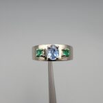 ​A premium handcrafted men's 925 sterling silver ring featuring a large oval-cut natural Ceylon blue sapphire center stone flanked by two vibrant, square-cut Swat Valley emerald side stones, displayed on a clean reflective surface.