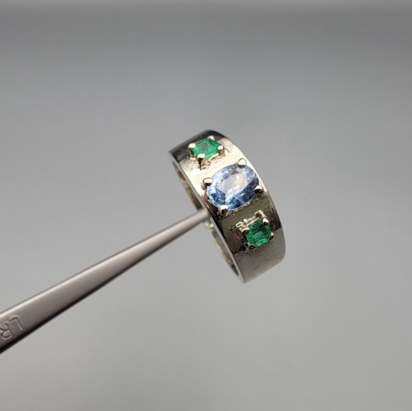 ​A premium handcrafted men's 925 sterling silver ring featuring a large oval-cut natural Ceylon blue sapphire center stone flanked by two vibrant, square-cut Swat Valley emerald side stones, displayed on a clean reflective surface.