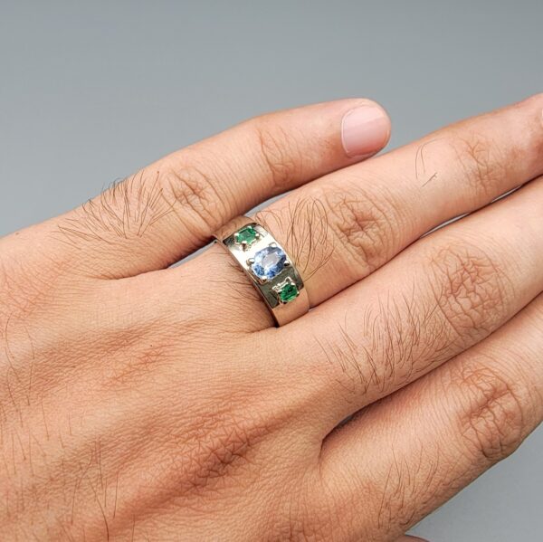 ​A premium handcrafted men's 925 sterling silver ring featuring a large oval-cut natural Ceylon blue sapphire center stone flanked by two vibrant, square-cut Swat Valley emerald side stones, displayed on a clean reflective surface.