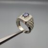 ​A premium men's 925 sterling silver statement ring featuring an oval-cut natural Ceylon blue sapphire centerpiece surrounded by a brilliant double border of sparkling white zircons, displayed from multiple professional angles on a reflective surface.