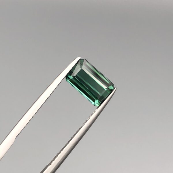 A natural 2.63-carat greenish-blue tourmaline from Africa, displaying a premium faceted rectangle emerald cut with deep color saturation and flawless transparency, held firmly in professional jeweler's tweezers.