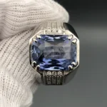 A luxury men's 925 sterling silver statement ring featuring a massive 11.80-carat oval-cut natural Ceylon blue sapphire surrounded by a brilliant halo of 0.47 carats of white diamonds, resting inside a textured white leather jewelry box liner.