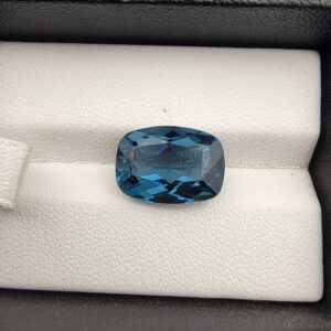​A premium 7.94-carat natural London Blue Topaz gemstone resting on a white textured leather insert inside a black display box. The stone is precision cut into an elegant cushion shape, exhibiting a deep, saturated ink-blue body with dazzling, brilliant secondary blue flashes across its expertly faceted crown.