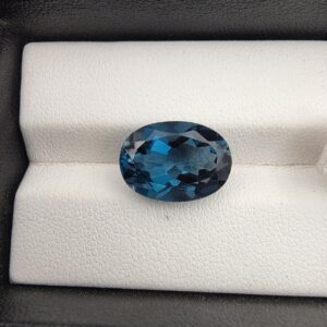 ​A premium 8.03-carat natural London Blue Topaz gemstone resting on a white textured leather insert inside a black display box. The stone is precision cut into an elegant oval shape, exhibiting a deep, saturated ink-blue body with dazzling, brilliant secondary blue flashes across its expertly faceted crown.