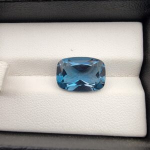 A premium 7.47-carat natural London Blue Topaz gemstone resting on a white textured leather insert inside a black display box. The stone is precision cut into an elegant cushion shape, exhibiting a deep, saturated ink-blue body with dazzling, brilliant secondary blue flashes across its expertly faceted crown.