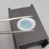 A premium 7-carat oval natural Ethiopian opal held in tweezers. Under standard light, it shows a translucent body with subtle neon blue and green flashes, while backlighting reveals a brilliant, fiery matrix of electric green, orange, and golden spark patterns.