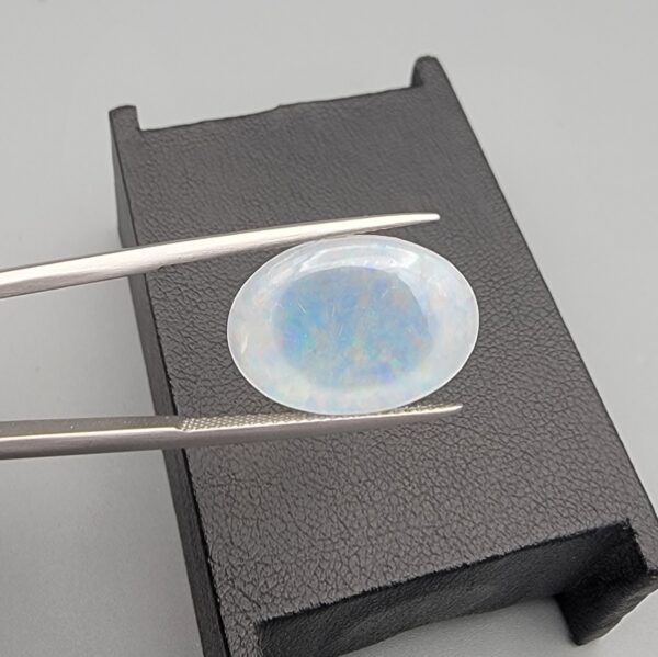 A premium 7-carat oval natural Ethiopian opal held in tweezers. Under standard light, it shows a translucent body with subtle neon blue and green flashes, while backlighting reveals a brilliant, fiery matrix of electric green, orange, and golden spark patterns.