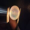 A premium 7-carat oval natural Ethiopian opal held in tweezers. Under standard light, it shows a translucent body with subtle neon blue and green flashes, while backlighting reveals a brilliant, fiery matrix of electric green, orange, and golden spark patterns.