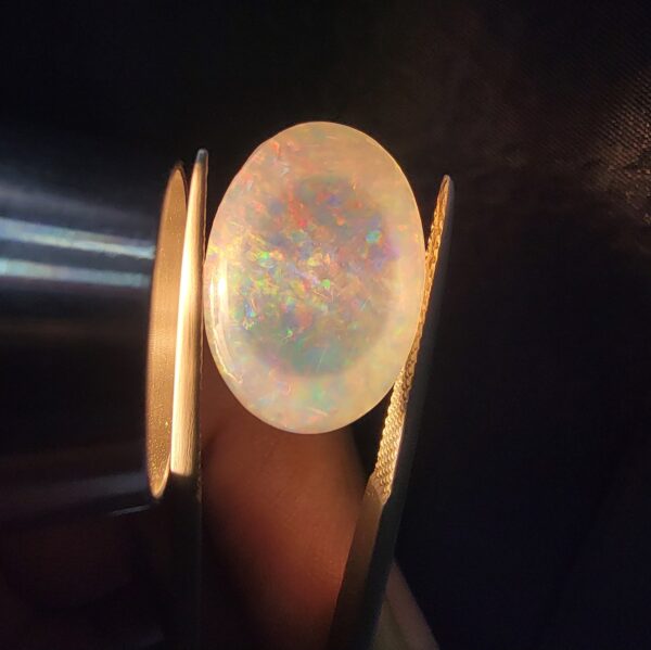 A premium 7-carat oval natural Ethiopian opal held in tweezers. Under standard light, it shows a translucent body with subtle neon blue and green flashes, while backlighting reveals a brilliant, fiery matrix of electric green, orange, and golden spark patterns.