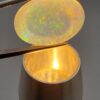 A premium 7-carat oval natural Ethiopian opal held in tweezers. Under standard light, it shows a translucent body with subtle neon blue and green flashes, while backlighting reveals a brilliant, fiery matrix of electric green, orange, and golden spark patterns.