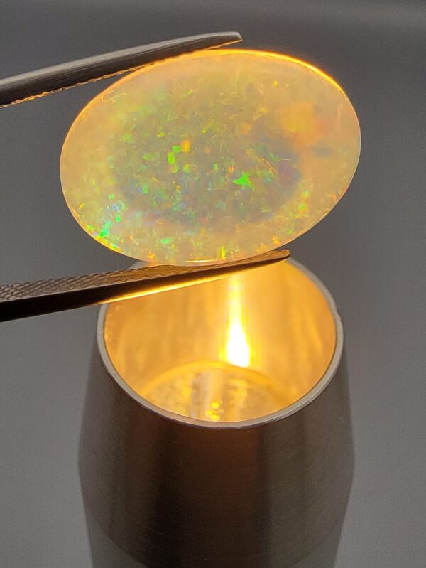 A premium 7-carat oval natural Ethiopian opal held in tweezers. Under standard light, it shows a translucent body with subtle neon blue and green flashes, while backlighting reveals a brilliant, fiery matrix of electric green, orange, and golden spark patterns.
