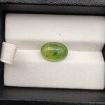 ​A rare 4.70-carat natural Italian Vesuvianite cabochon resting on a white textured leather insert inside a black display box. The stone exhibits a smooth, high-dome oval cut with excellent symmetry, showcasing a vibrant, translucent yellow-green to chartreuse body with distinctive, deeper green mottled inclusions.