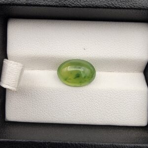 ​A rare 4.70-carat natural Italian Vesuvianite cabochon resting on a white textured leather insert inside a black display box. The stone exhibits a smooth, high-dome oval cut with excellent symmetry, showcasing a vibrant, translucent yellow-green to chartreuse body with distinctive, deeper green mottled inclusions.