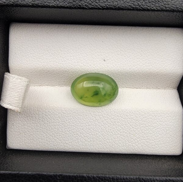 ​A rare 4.70-carat natural Italian Vesuvianite cabochon resting on a white textured leather insert inside a black display box. The stone exhibits a smooth, high-dome oval cut with excellent symmetry, showcasing a vibrant, translucent yellow-green to chartreuse body with distinctive, deeper green mottled inclusions.