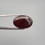 ​A massive 22.20-carat oval natural Yemeni carnelian agate cabochon held in tweezers. Without direct light, the gemstone displays an opaque, deep liver-brown tone, but under direct backlighting, it transforms into a fully translucent, deep crimson-red stone showing faint internal agate structure.