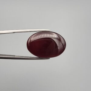 ​A massive 22.20-carat oval natural Yemeni carnelian agate cabochon held in tweezers. Without direct light, the gemstone displays an opaque, deep liver-brown tone, but under direct backlighting, it transforms into a fully translucent, deep crimson-red stone showing faint internal agate structure.