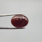 A massive 21.58-carat oval natural Yemeni carnelian agate cabochon held securely in tweezers. In normal lighting as seen in input_file_0.png, the stone shows a smooth, glossy, deep liver-brown body. Under strong direct backlighting in input_file_1.png and input_file_2.png, it glows with a rich, uniform translucent reddish-crimson color, revealing elegant natural parallel banding lines along its lower edge.