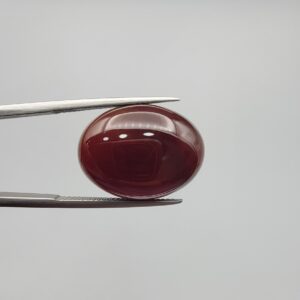 A massive 21.58-carat oval natural Yemeni carnelian agate cabochon held securely in tweezers. In normal lighting as seen in input_file_0.png, the stone shows a smooth, glossy, deep liver-brown body. Under strong direct backlighting in input_file_1.png and input_file_2.png, it glows with a rich, uniform translucent reddish-crimson color, revealing elegant natural parallel banding lines along its lower edge.