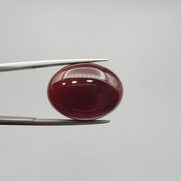 A massive 21.58-carat oval natural Yemeni carnelian agate cabochon held securely in tweezers. In normal lighting as seen in input_file_0.png, the stone shows a smooth, glossy, deep liver-brown body. Under strong direct backlighting in input_file_1.png and input_file_2.png, it glows with a rich, uniform translucent reddish-crimson color, revealing elegant natural parallel banding lines along its lower edge.