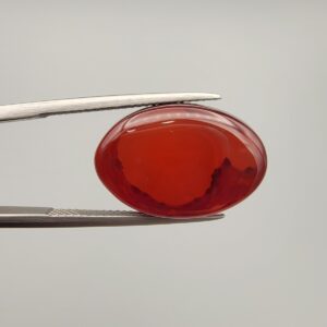 ​A premium 14.26-carat natural oval Yemeni carnelian agate cabochon held in tweezers. While appearing dark brown with a dense core in flat shadow, strong backlighting transforms it into a brilliant, translucent fiery orange gemstone displaying elegant, natural dendritic cloud-like agate zoning patterns along its lower and side edges.