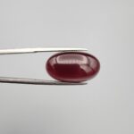 A premium 19.00-carat oval natural Yemeni carnelian agate cabochon held securely in tweezers. In normal lighting as seen in the first image, the stone shows a smooth, glossy, deep liver-brown body with a mirror-like finish. Under strong direct backlighting in the second image, it glows with a rich, uniform translucent reddish-crimson color across its entire surface.