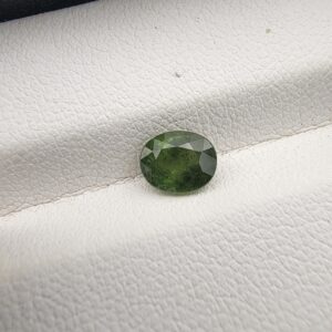 A premium 1.20-carat natural Madagascar Green Sapphire gemstone resting inside a black presentation box on a white textured leather insert. The loose corundum gem is precision faceted into a beautifully symmetrical oval shape, exhibiting a rich forest-green to olive body color with sharp, light-reflecting crown facets.