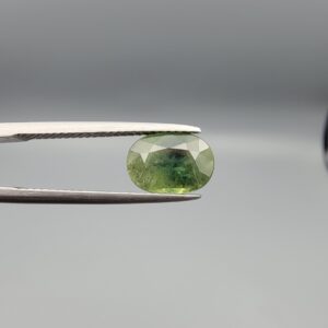 A premium 3.09-carat natural Madagascar Green Sapphire gemstone held securely in silver metal tweezers against a clean gray background. The loose corundum gem is precision faceted into a beautifully symmetrical oval shape, exhibiting a bright mint-green body with a concentrated teal-green center core and sharp, light-reflecting crown facets.