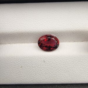​A premium 1.64-carat natural Mozambique Rubellite Tourmaline gemstone. In the first image, it is held securely in silver metal tweezers against a gray backdrop, flashing a vibrant magenta-red under direct light. The second image shows the loose oval faceted gemstone resting flat on a white textured leather insert inside a display box, showcasing its rich, deep crimson body color.